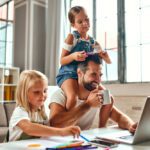 A father works on a laptop while his daughters play.