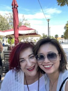 Two women smiling outdoors under an umbrella.